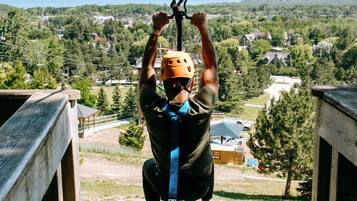 A person in a helmet and harness suspended by a rope, with a view of a wooded area and buildings in the background.