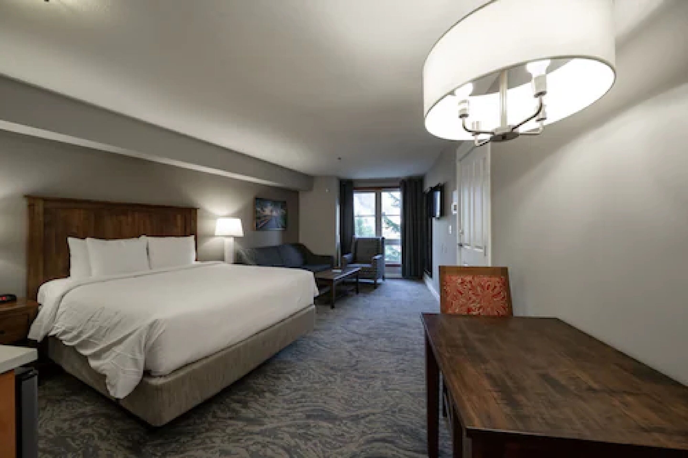 A hotel room with a large bed, a desk, a chair, a lamp, and a view of the outdoors.