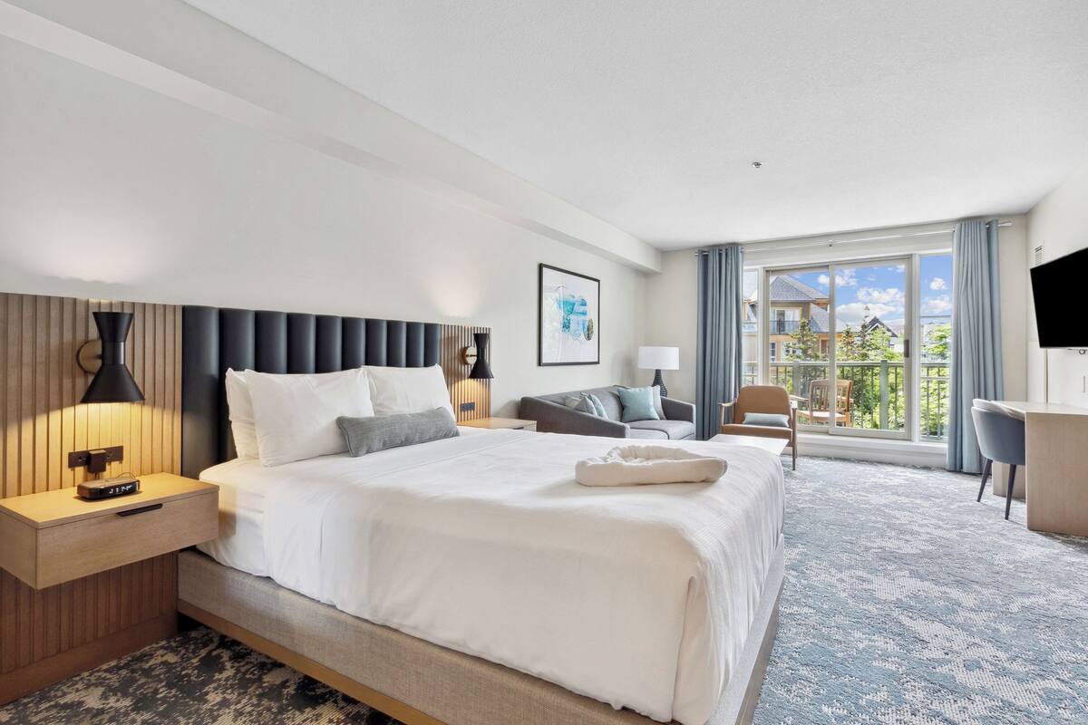 A modern hotel room with a large bed, a desk, a chair, and a balcony with a view.