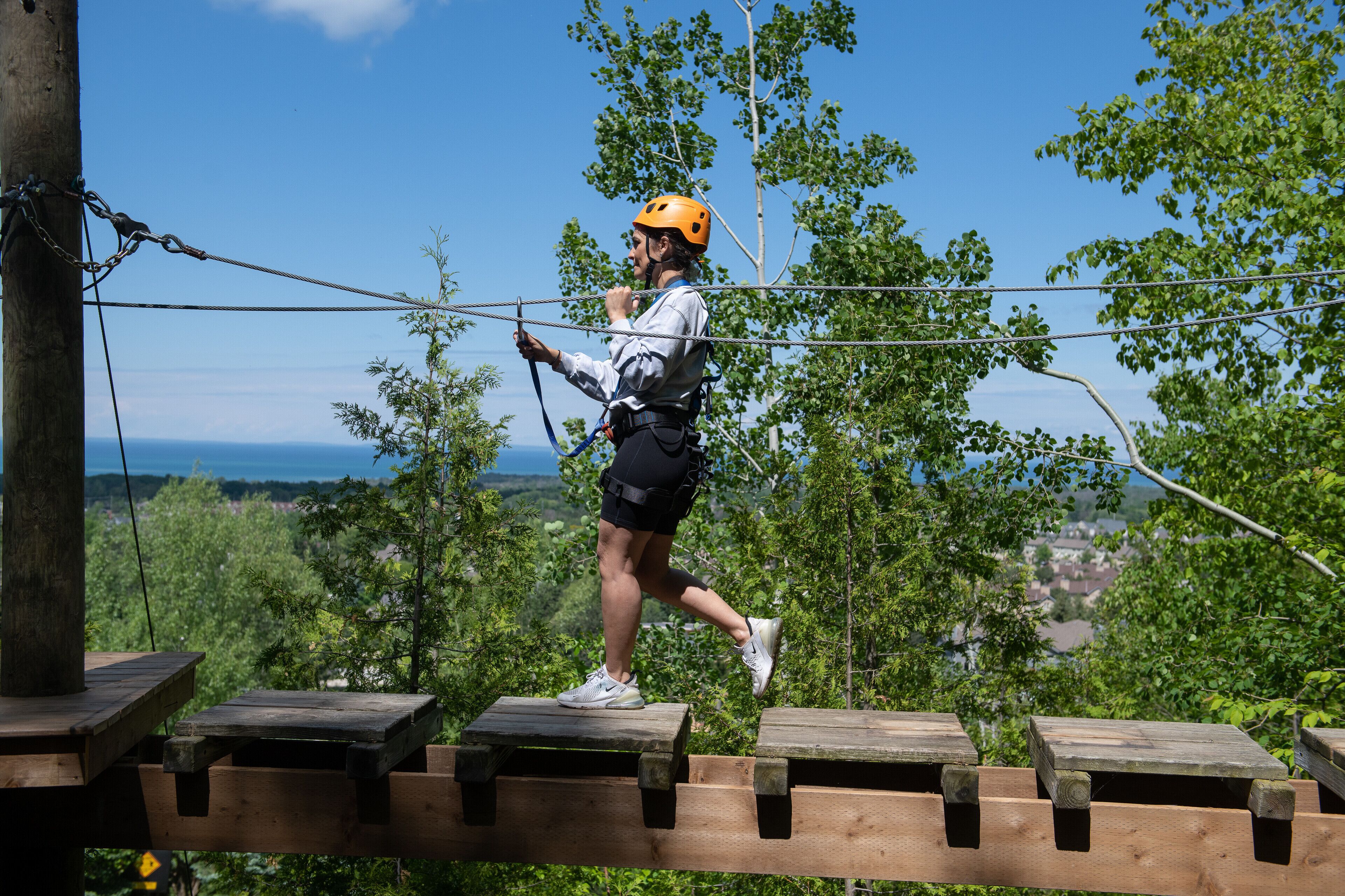 A person navigating a rope bridge in a natural setting with trees and a distant view of the landscape.