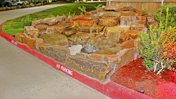 A landscaped area with a rock waterfall feature, a red curb indicating no parking, and a small tree. - Palacio Royale Inn Houston NW Beltway 8 (Houston)