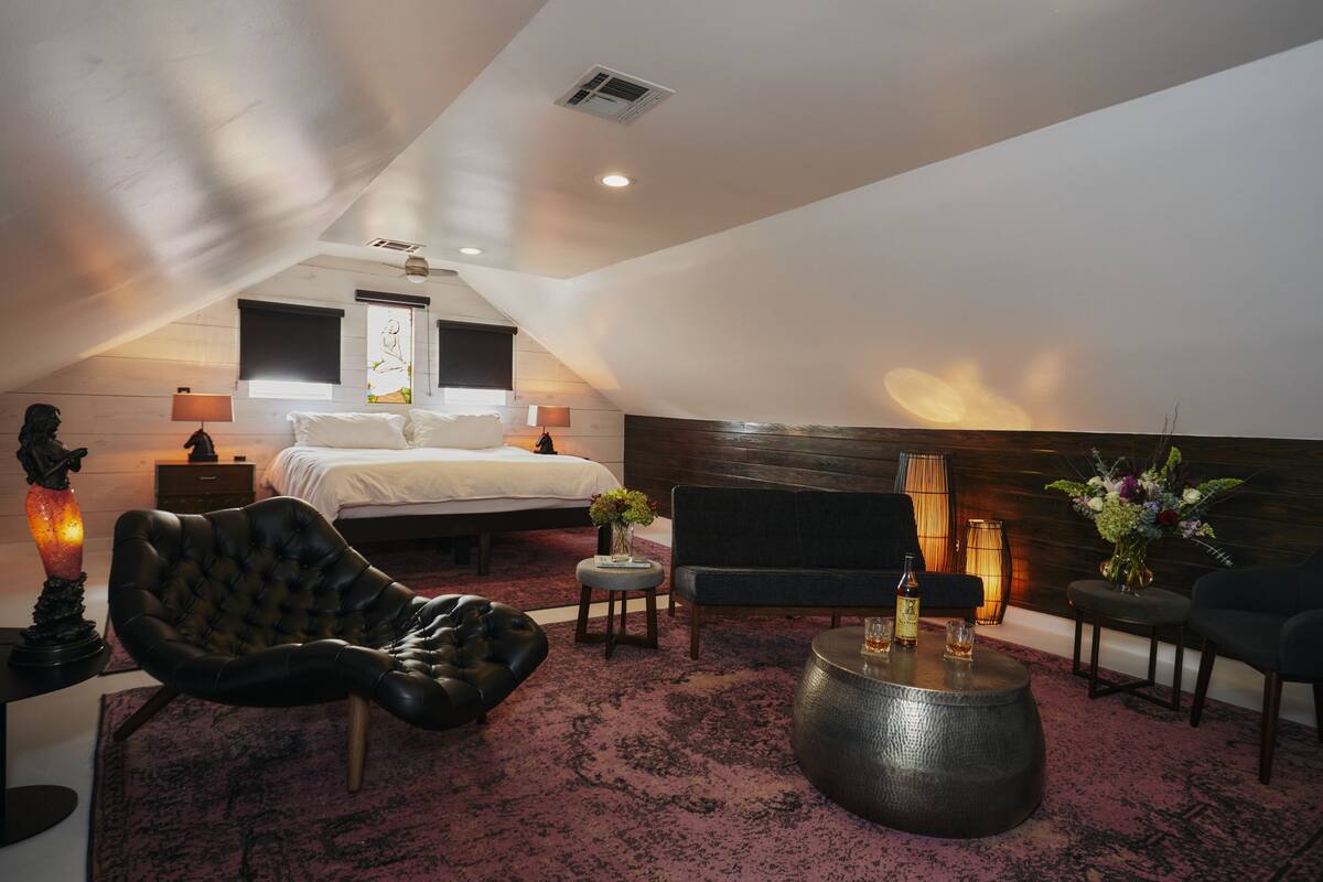 A modern bedroom with a large bed, a sitting area with a sofa and armchair, a coffee table, and a decorative vase.