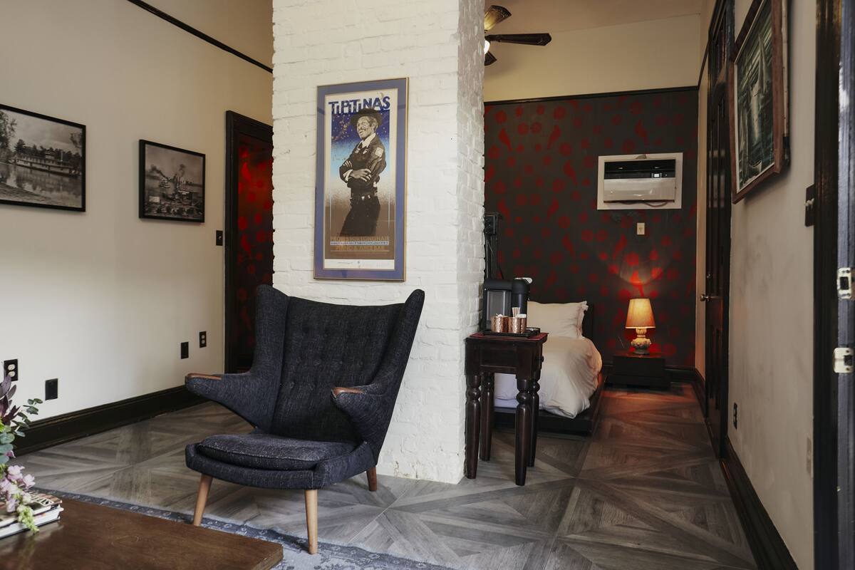 A hotel lobby with a dark armchair, a bed with a white pillow, a poster on the wall, and a patterned floor.