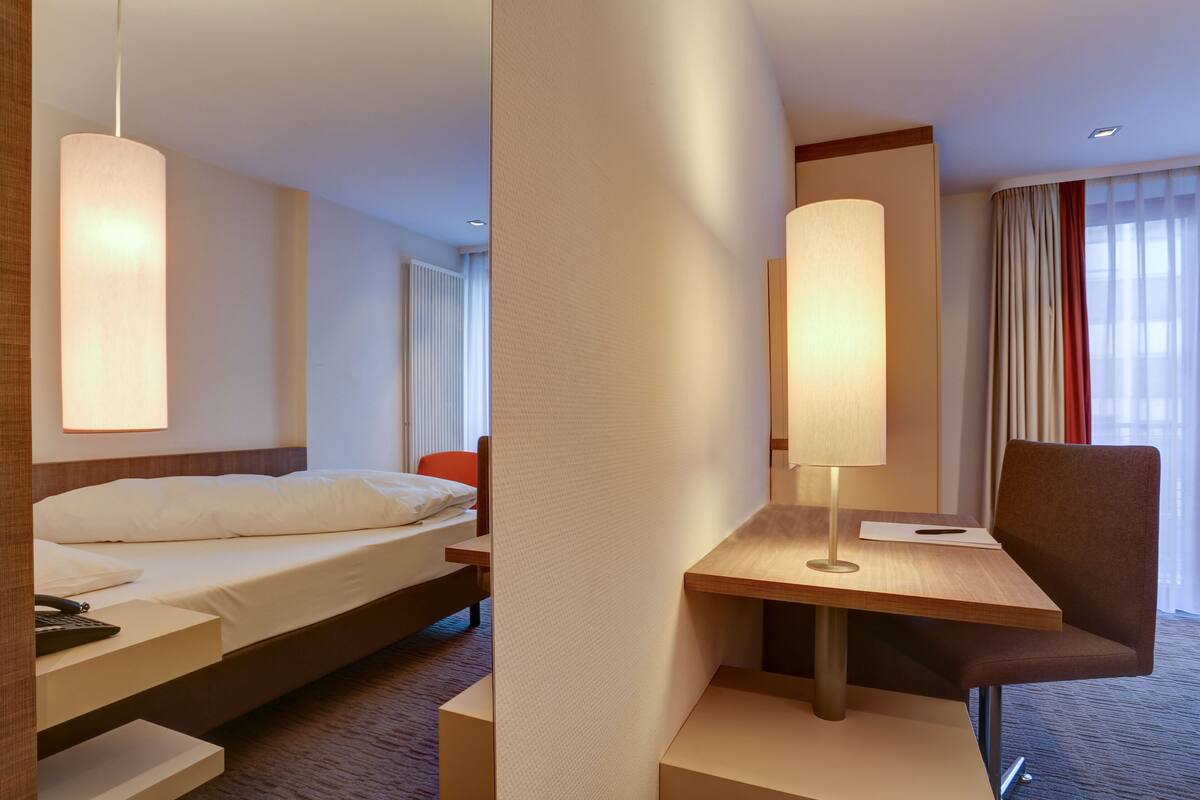 Una habitación de hotel con cama, escritorio con lámpara, silla y ventana con cortinas.