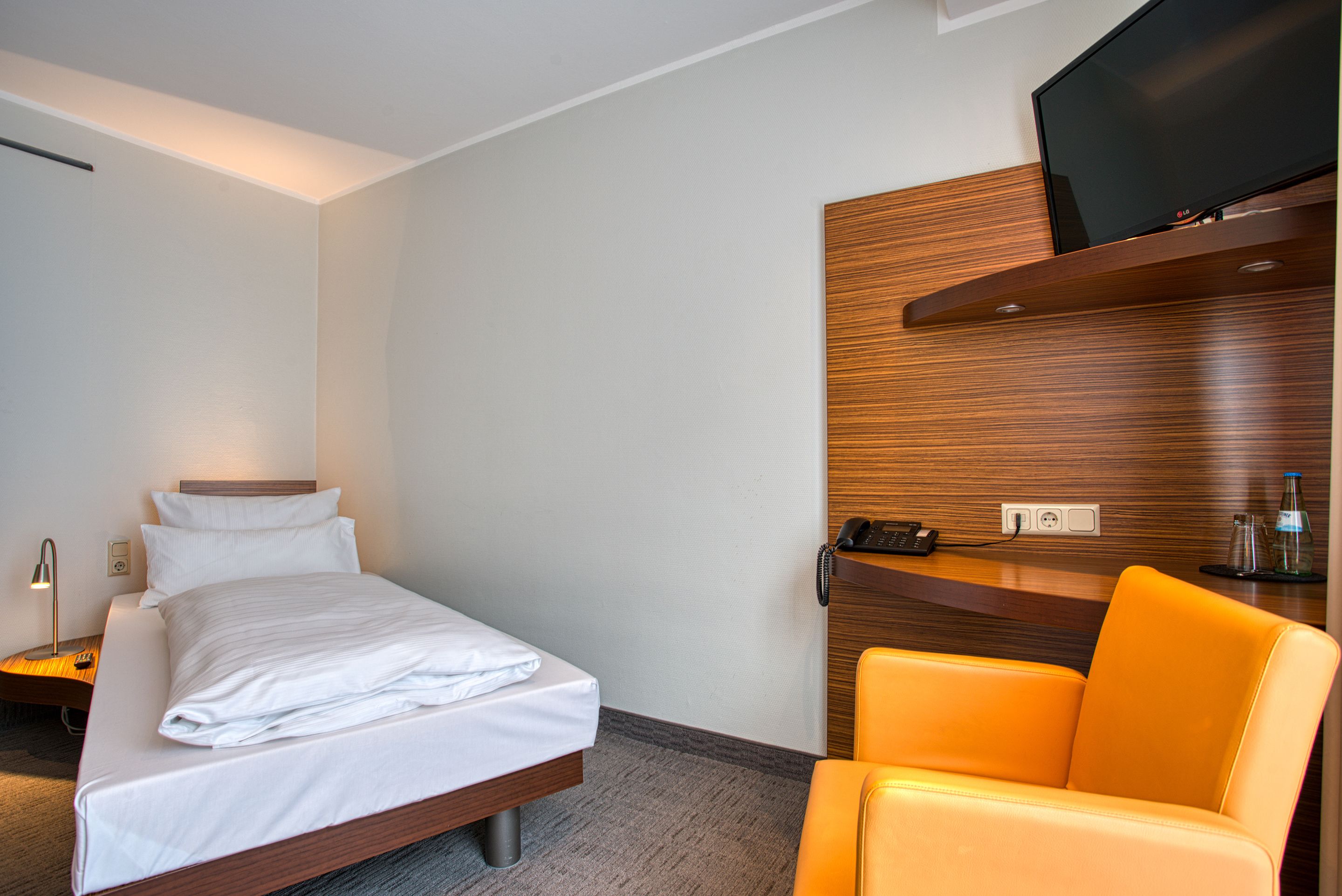 Habitación de hotel con cama, televisor montado en un panel de madera, escritorio con teléfono y una silla naranja.
