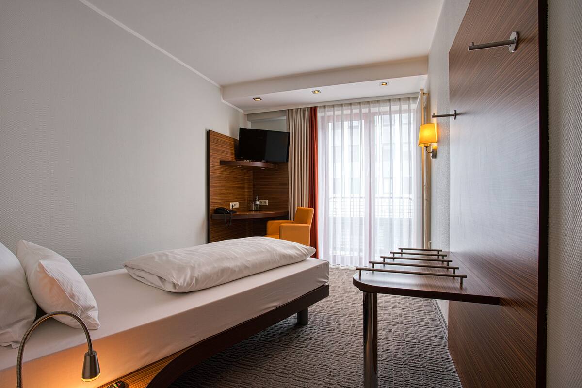 Habitación de hotel con una cama individual, una mesita, una silla, un televisor montado en la pared y una ventana con cortinas.