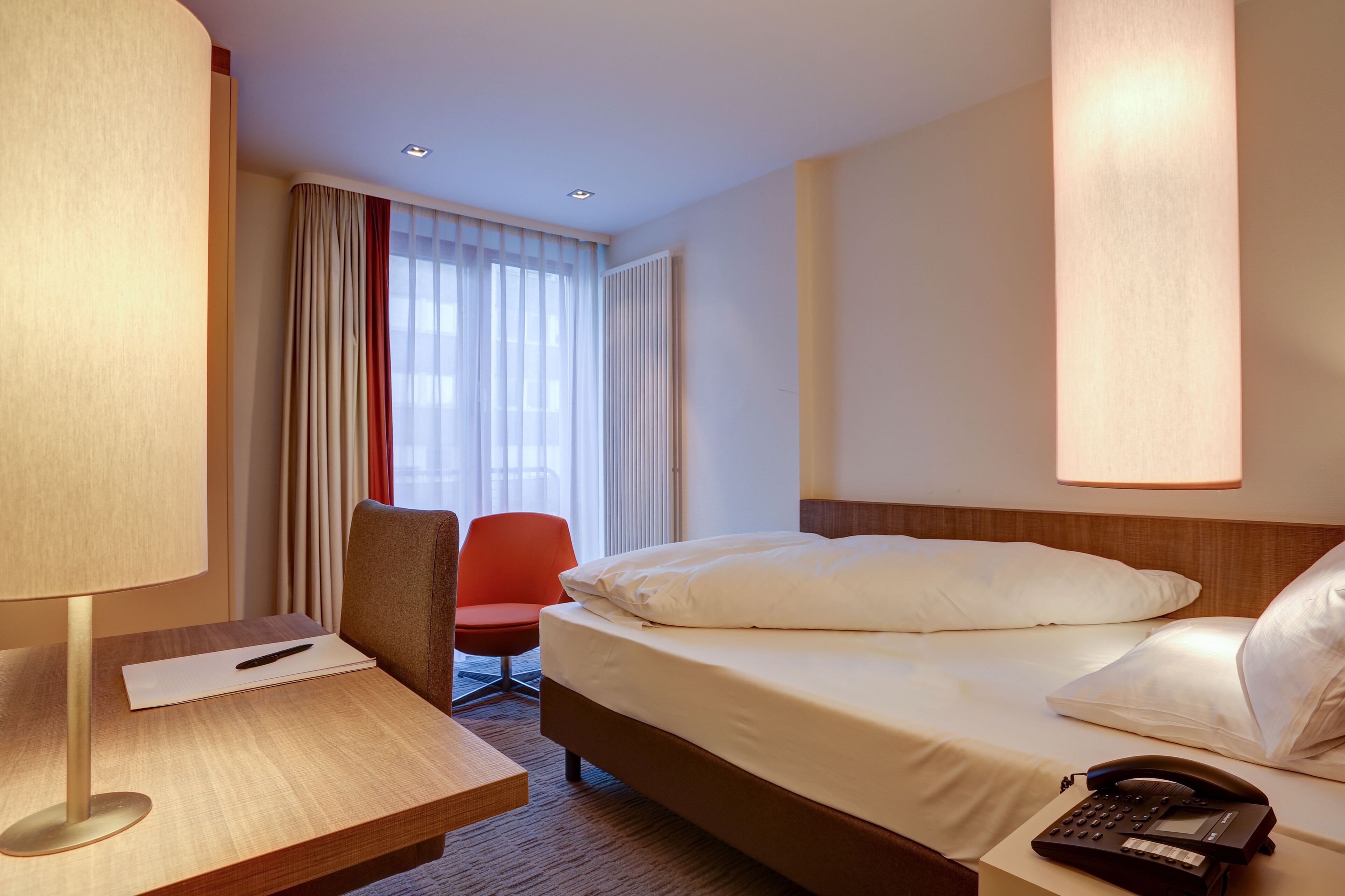 A hotel room with a bed, a desk with a lamp, a chair, and a telephone.