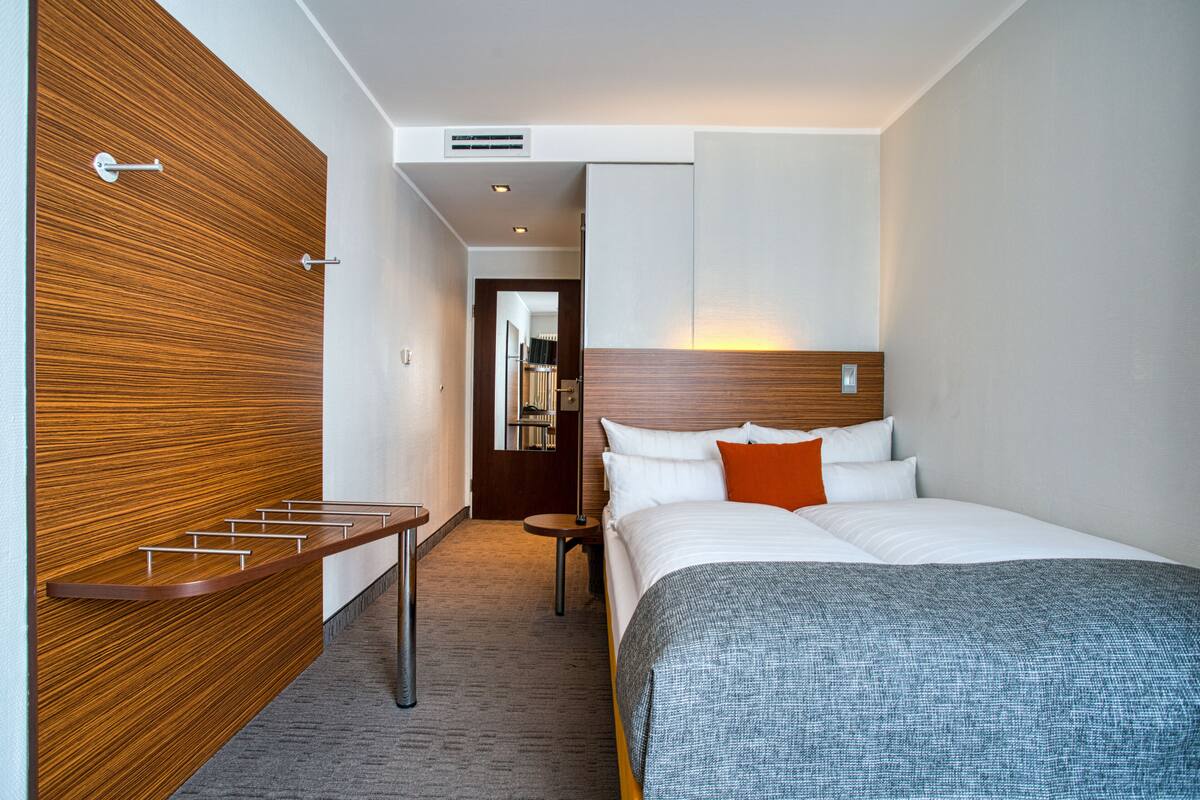 Una habitación de hotel moderna con una pared decorativa de madera, una cama bien hecha con almohadas blancas y anaranjadas, y una pequeña mesa redonda con una silla.