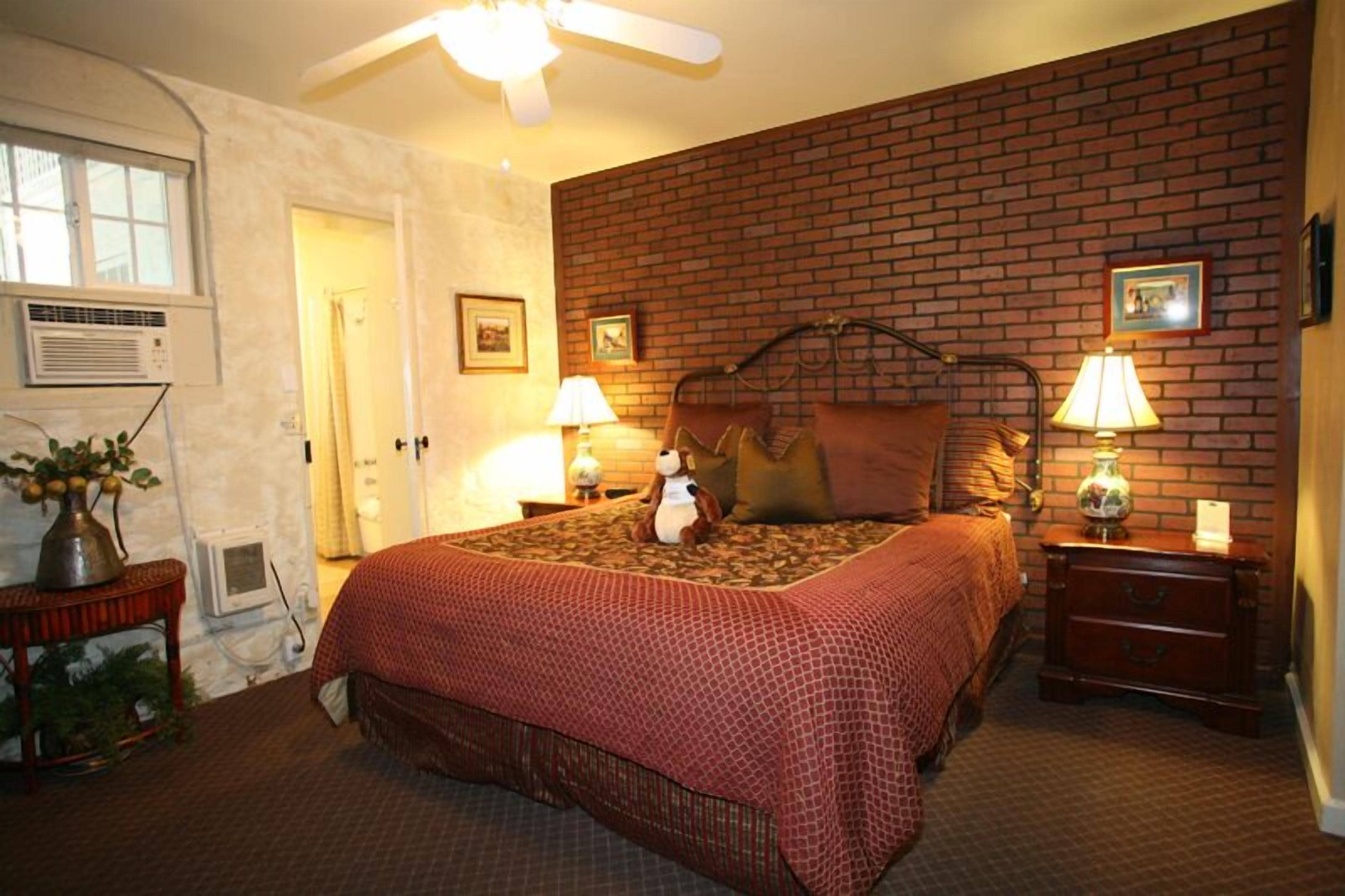 A bedroom with a brick-patterned headboard, two bedside lamps, a bed with a red patterned bedspread, a nightstand with a lamp, and a ceiling fan.