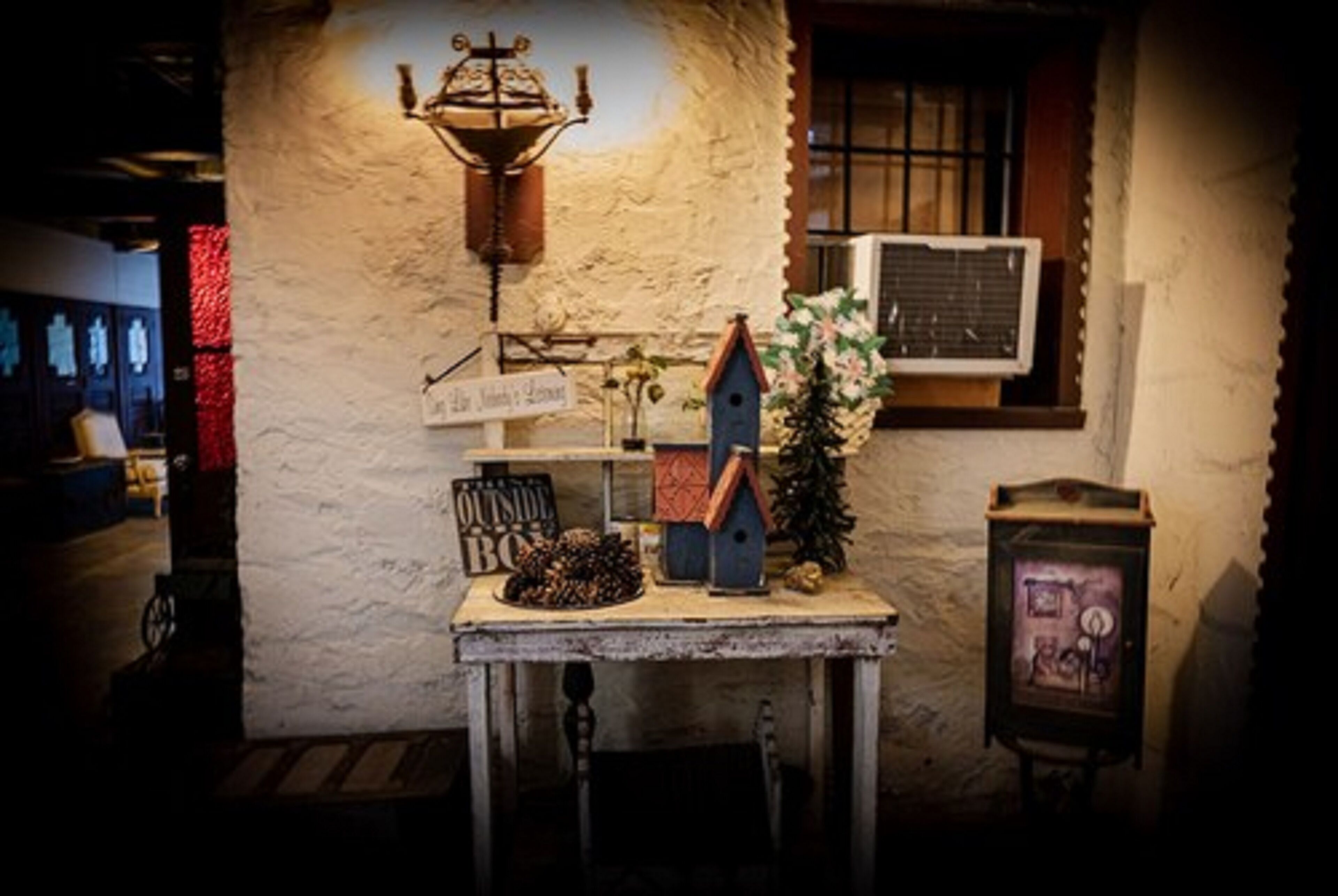 A rustic interior with a stone wall, a wooden table with a sign reading "OUTSIDE BO", a birdhouse, and a small Christmas tree.