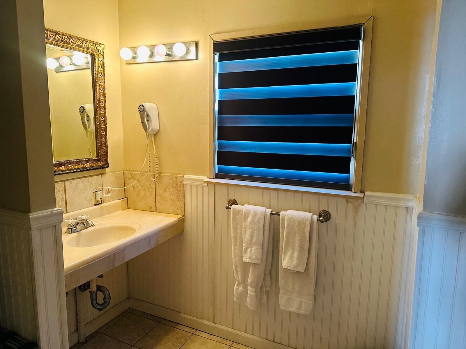 Un baño con lavamanos, tocador, espejo, secador de pelo y ventana con persianas azules.