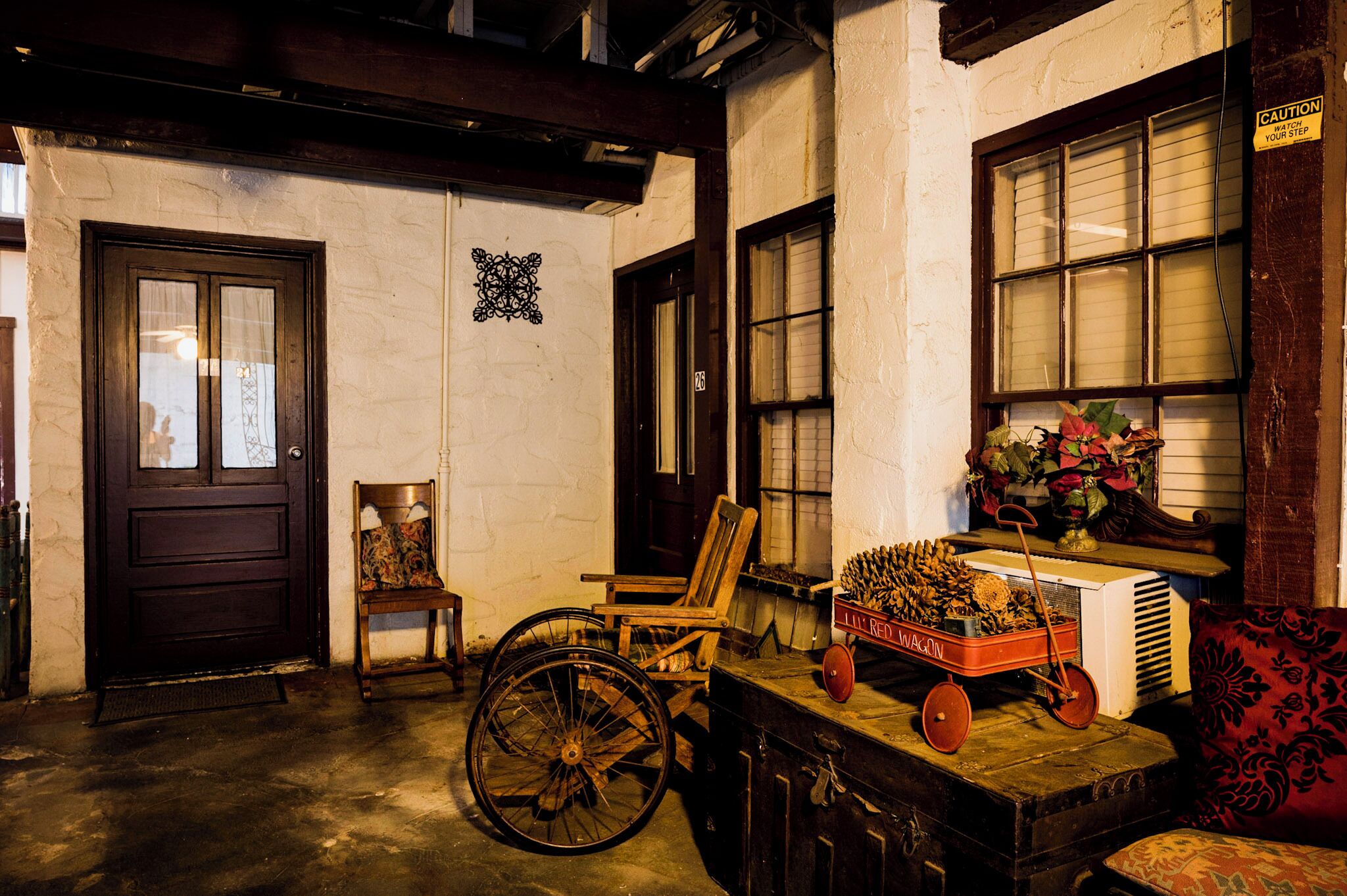 A room with a wooden door, a wheelchair, a vintage cart, and a red wagon wheel.