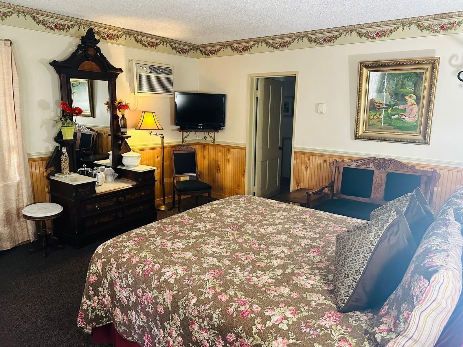 A bedroom with a floral bedspread, a dresser with a mirror, a television, and a painting on the wall.