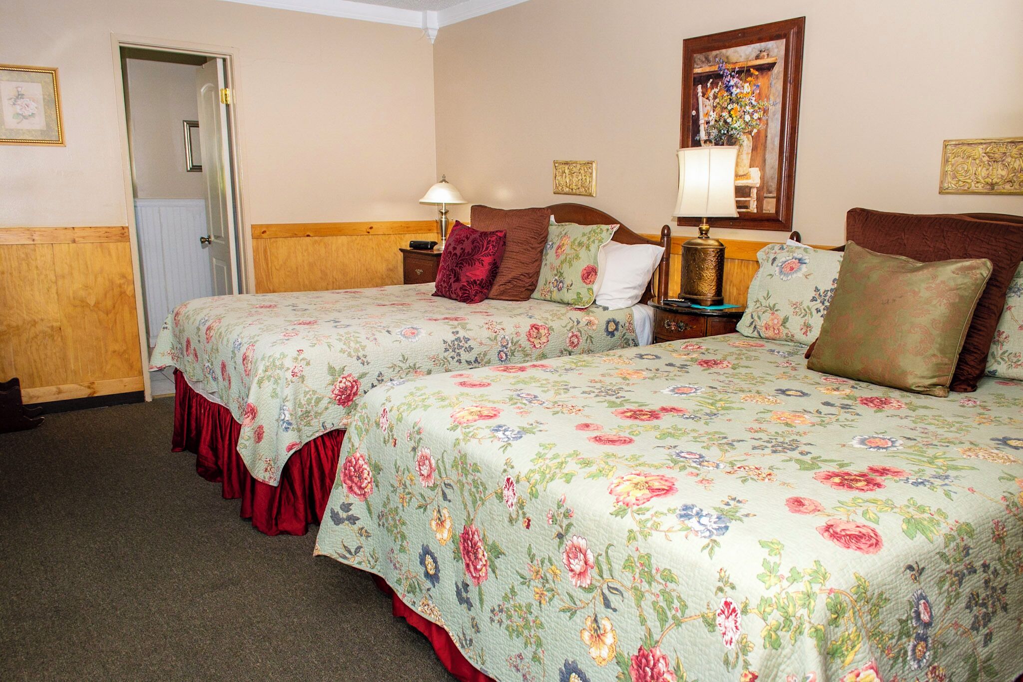 A hotel room with two beds, floral bedding, bedside tables with lamps, a door, and a mirror on the wall.