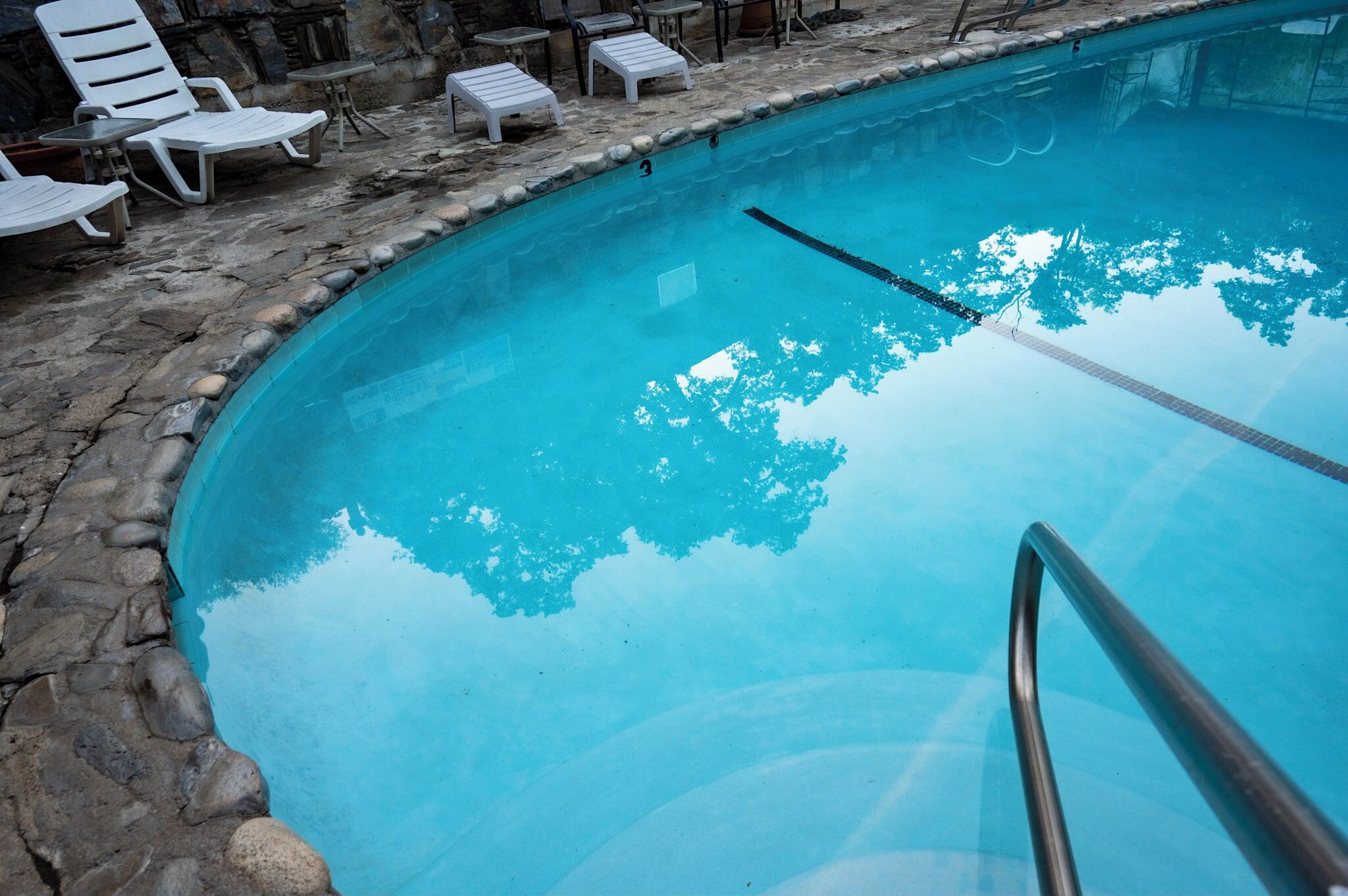 Una piscina con borde de piedra, sillas de descanso y barandilla de metal.