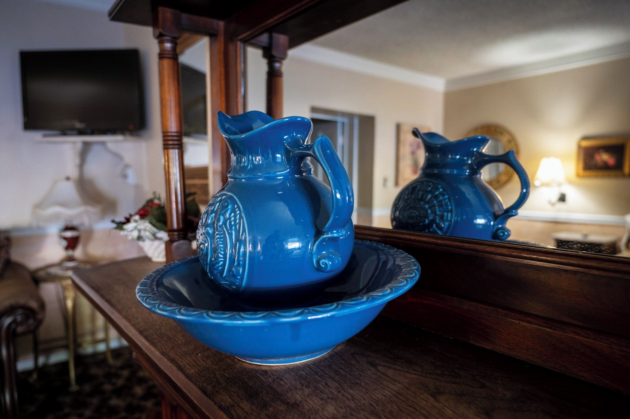A blue pitcher on a dark wooden tray, with a mirror reflecting the pitcher and a television in the background.