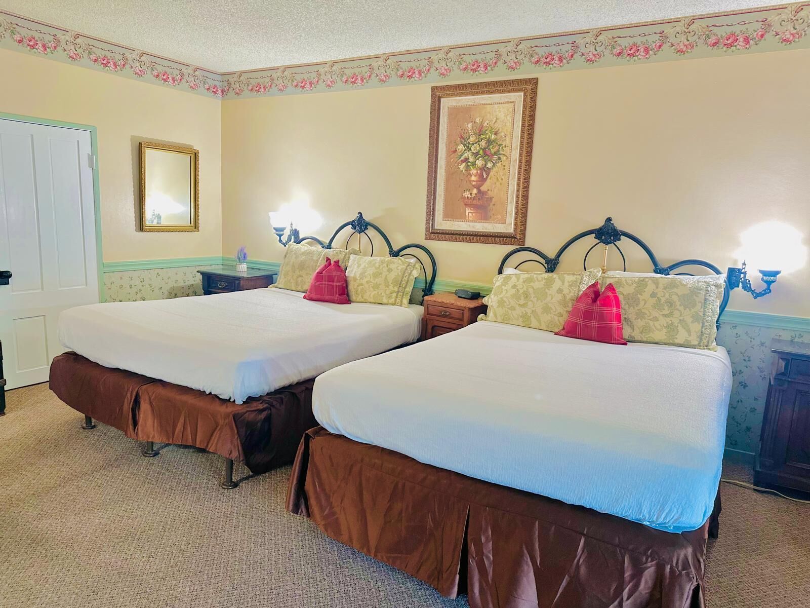 Habitación de hotel con dos camas, una puerta, una mesita de noche, una lámpara y un papel tapiz con estampado floral.