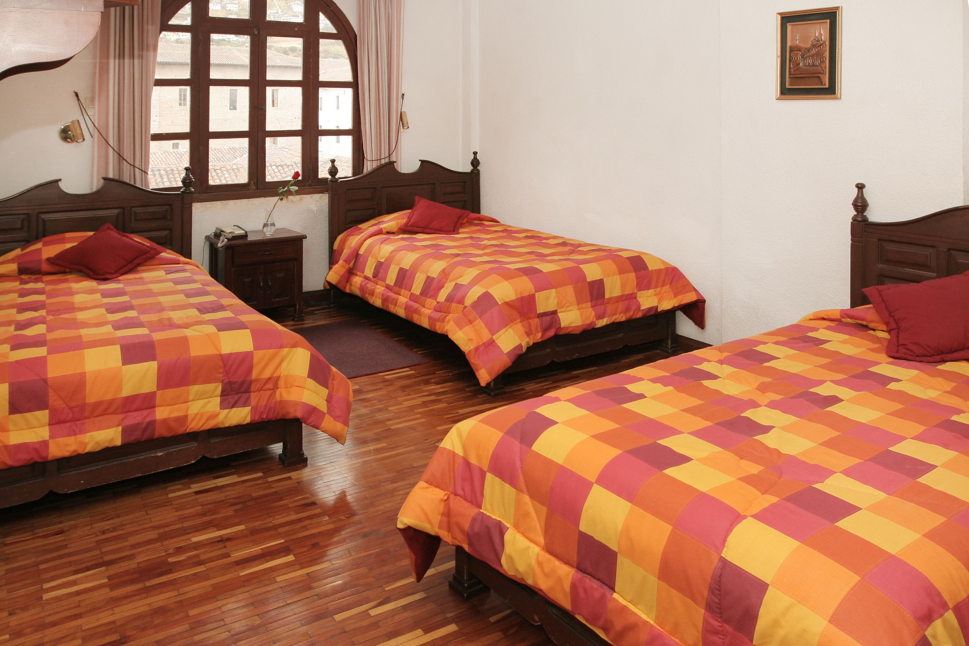 A room with three wooden beds, each with a checkered bedspread and pillows. There is a window with curtains, a small table with a vase, and a framed picture on the wall.