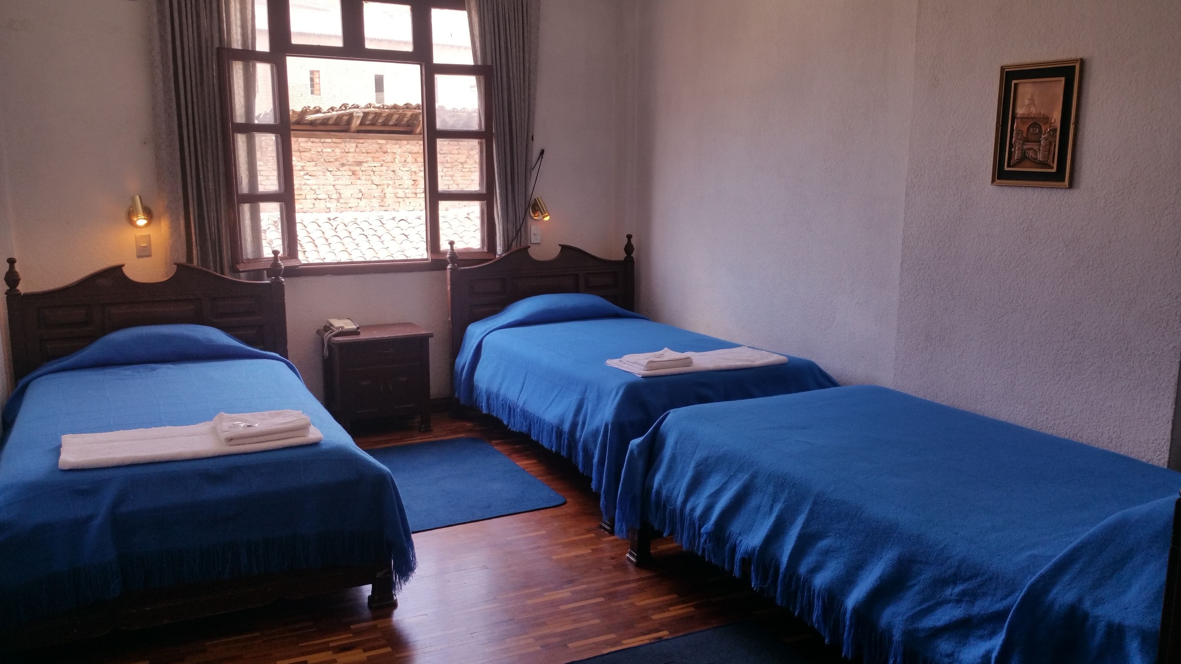 A room with two beds, wooden headboards, a small bedside table, a nightstand, a window with curtains, and a framed picture on the wall.