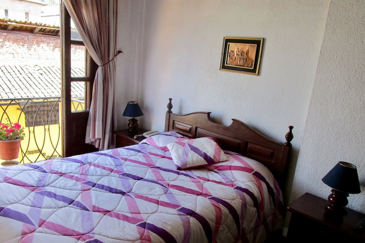 A neatly made bed with a patterned quilt, a wooden headboard, two bedside tables with lamps, a framed picture on the wall, and a balcony with a potted plant.