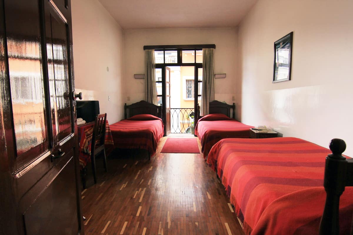 A hotel room with two single beds, a wooden floor, a red carpet, a balcony with a view, and a door with a mirror.
