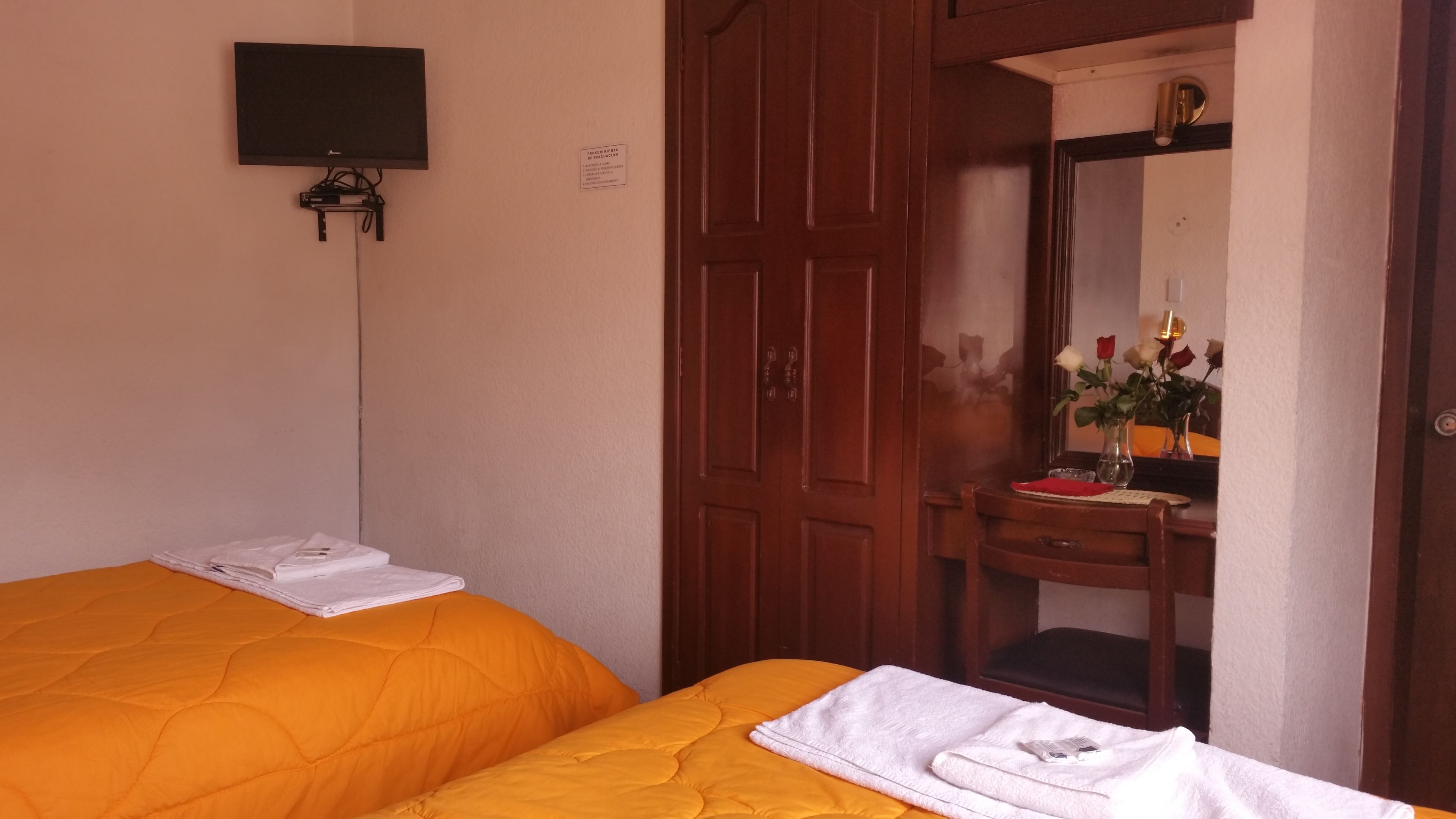 A hotel room with two beds, a TV mounted on the wall, a wooden wardrobe, a vanity with a mirror, and a vase with flowers.
