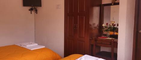 A hotel room with two beds, a TV mounted on the wall, a wooden wardrobe, a vanity with a mirror, and a vase with flowers.