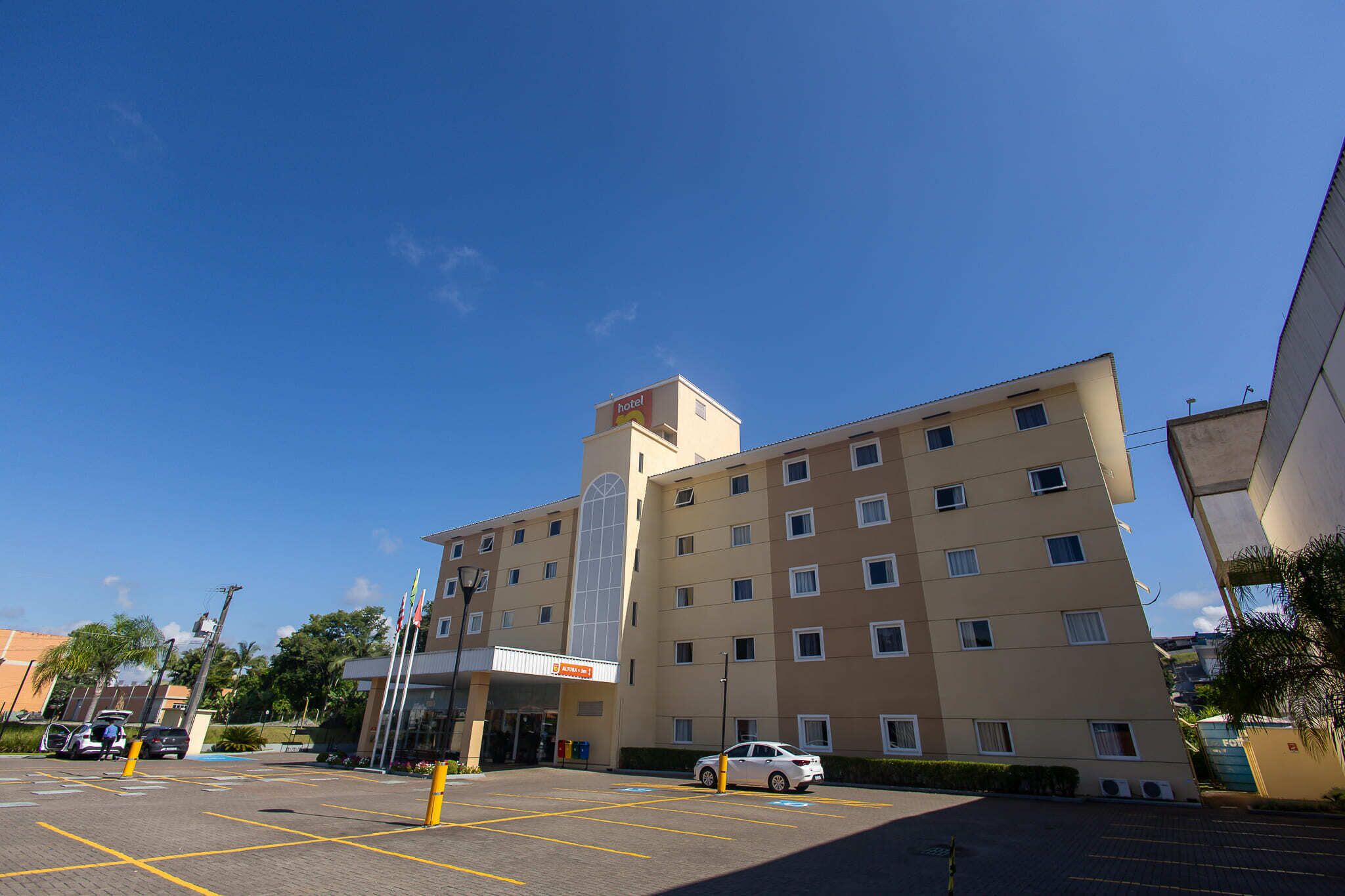 A multi-story hotel with a central entrance, a parking lot with marked spaces, and a clear blue sky.