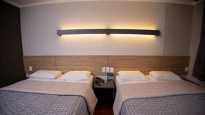 Habitación de hotel con dos camas, cabecero de madera, mesita de noche con un teléfono y un folleto, y una lámpara fijada a la pared.