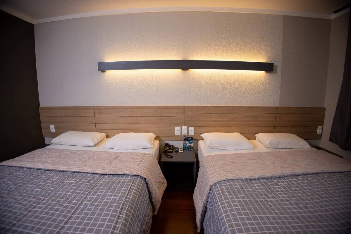 A hotel room with two beds, a wooden headboard, a nightstand with a phone and a brochure, and a wall-mounted light fixture.