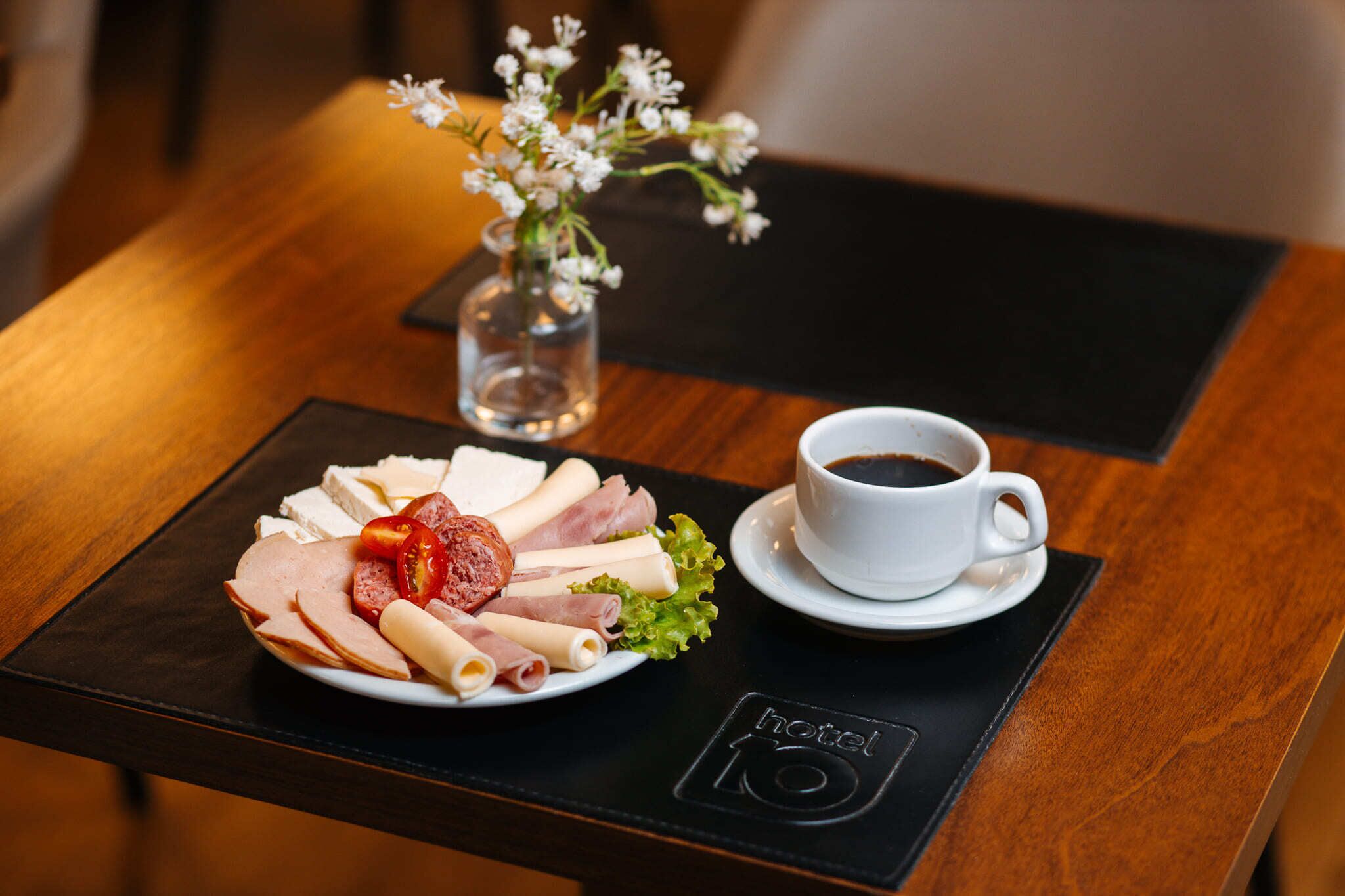 A breakfast tray with various meats, a cup of coffee, and a small vase with flowers on a wooden table.