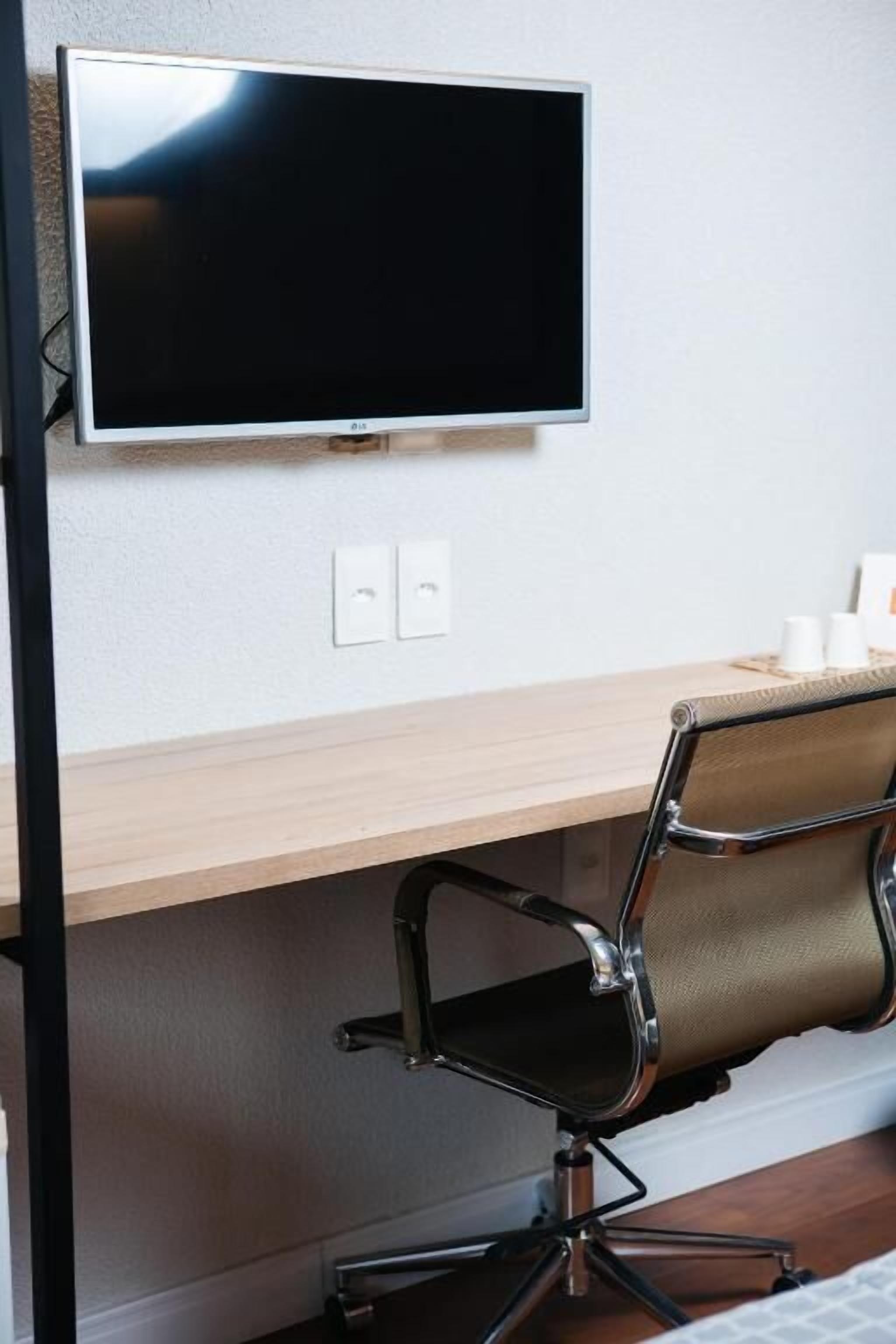 A modern office space with a flat-screen monitor mounted on the wall, a wooden desk, a black office chair, and a white wall with two electrical outlets.