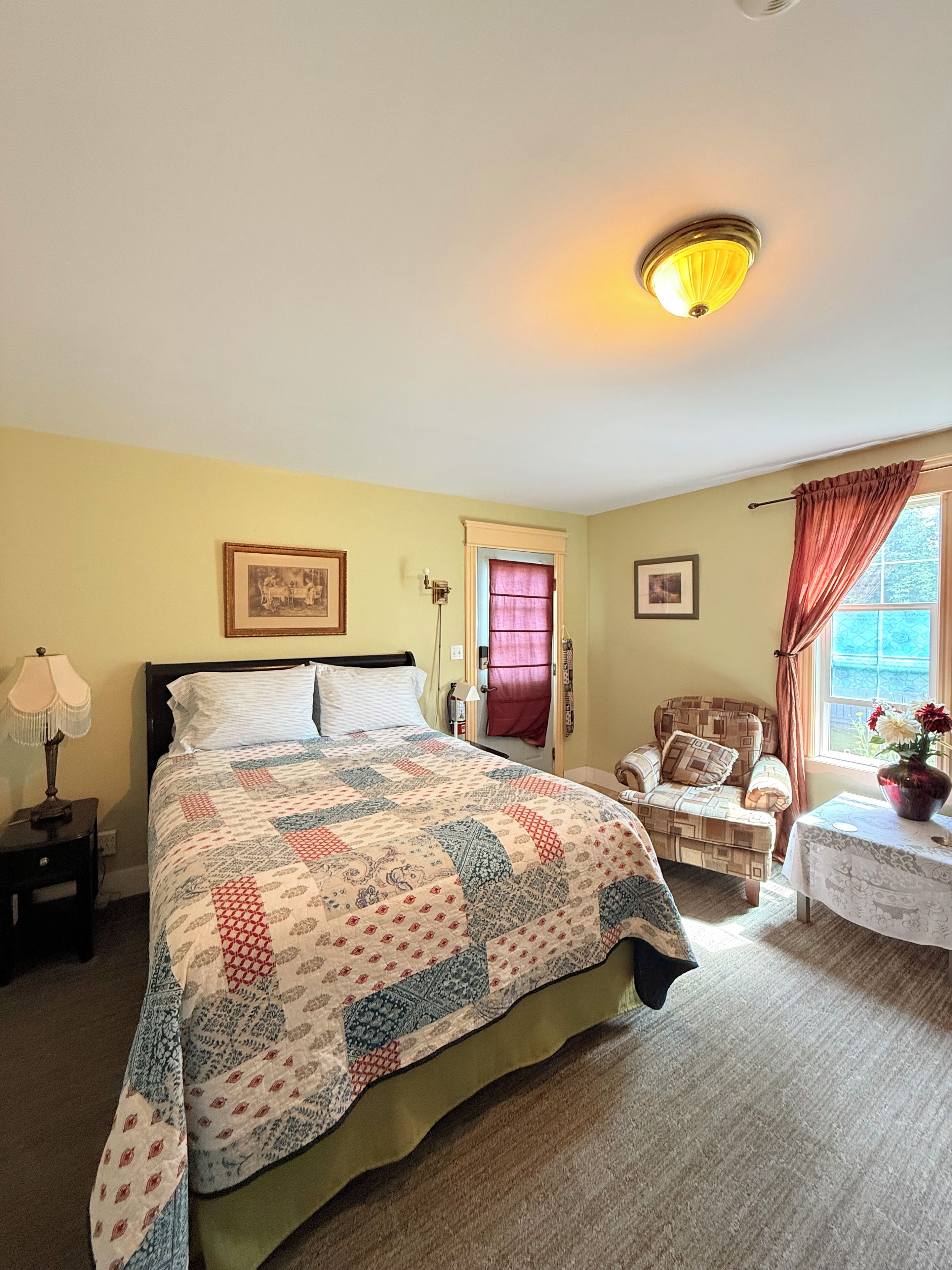 A bedroom with a bed, a chair, a table with a vase, and a window with curtains.