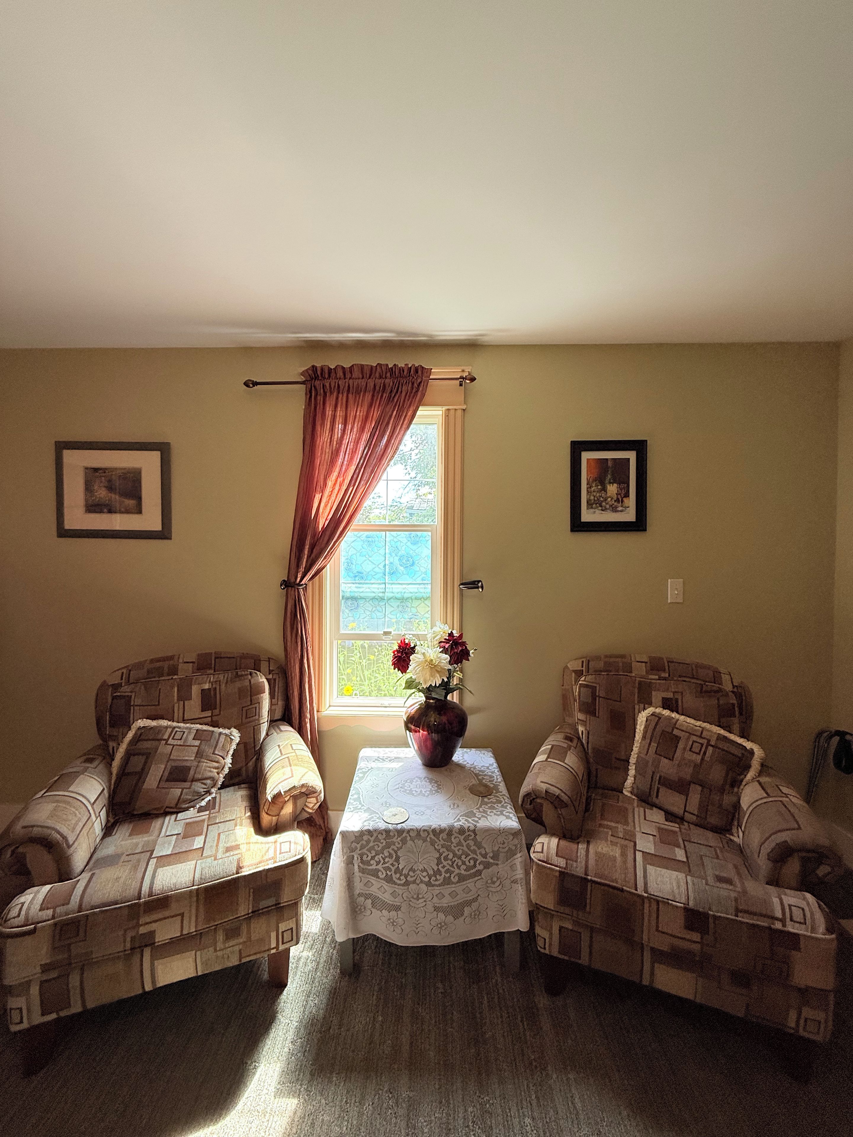 A room with two patterned armchairs, a small table with a vase of flowers, and a window with curtains.