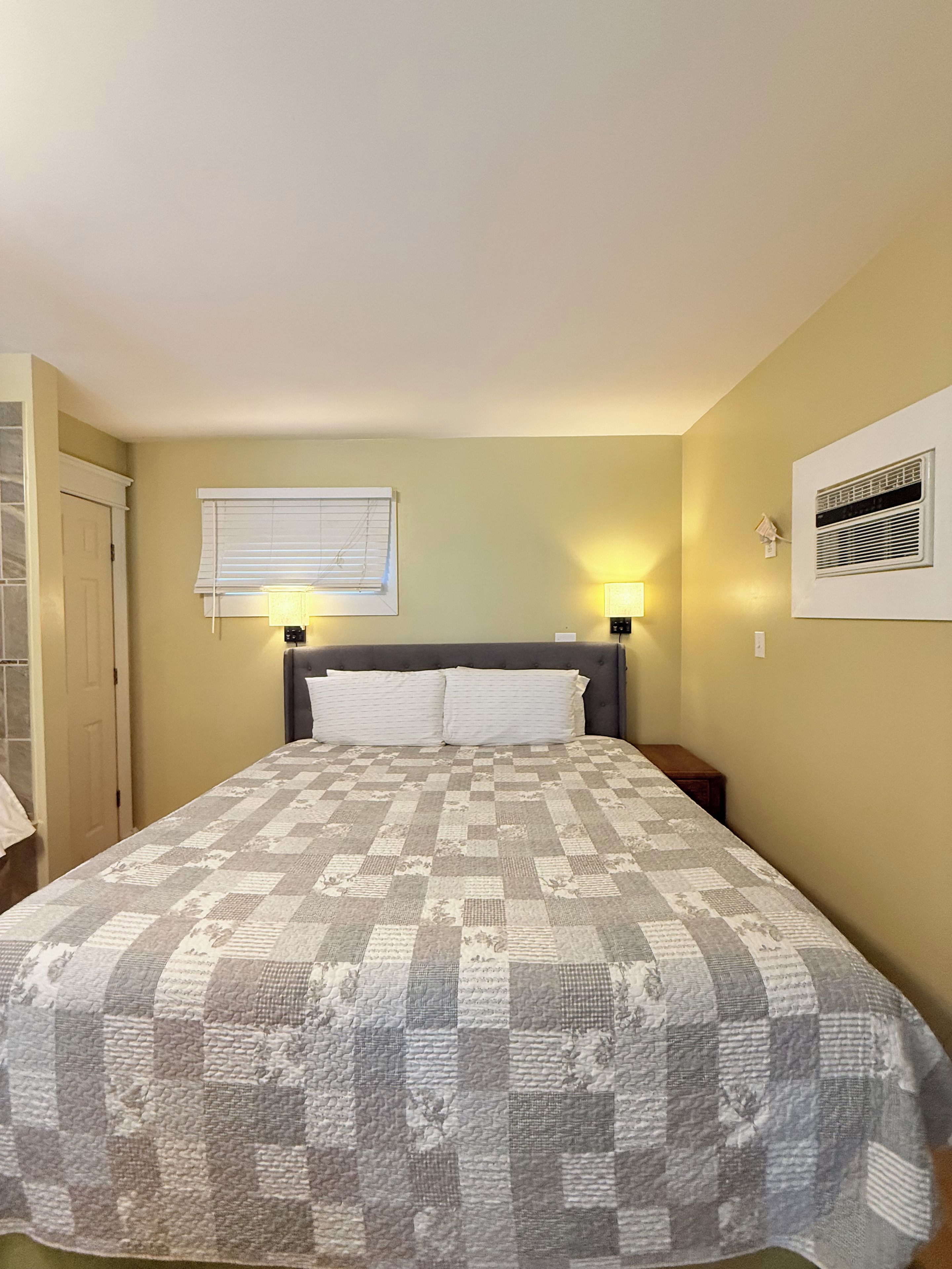 Un dormitorio con cama, dos lámparas de noche, una ventana con persianas y un equipo de aire acondicionado en la pared.
