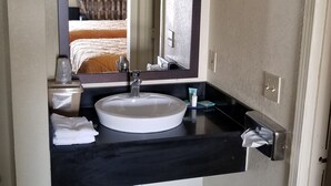Ein Badezimmer mit einem weißen Waschbecken, einer dunklen Arbeitsplatte und einem Spiegel darüber. Auf der Ablage liegt ein Handtuch und neben der Tür steht ein Mülleimer.