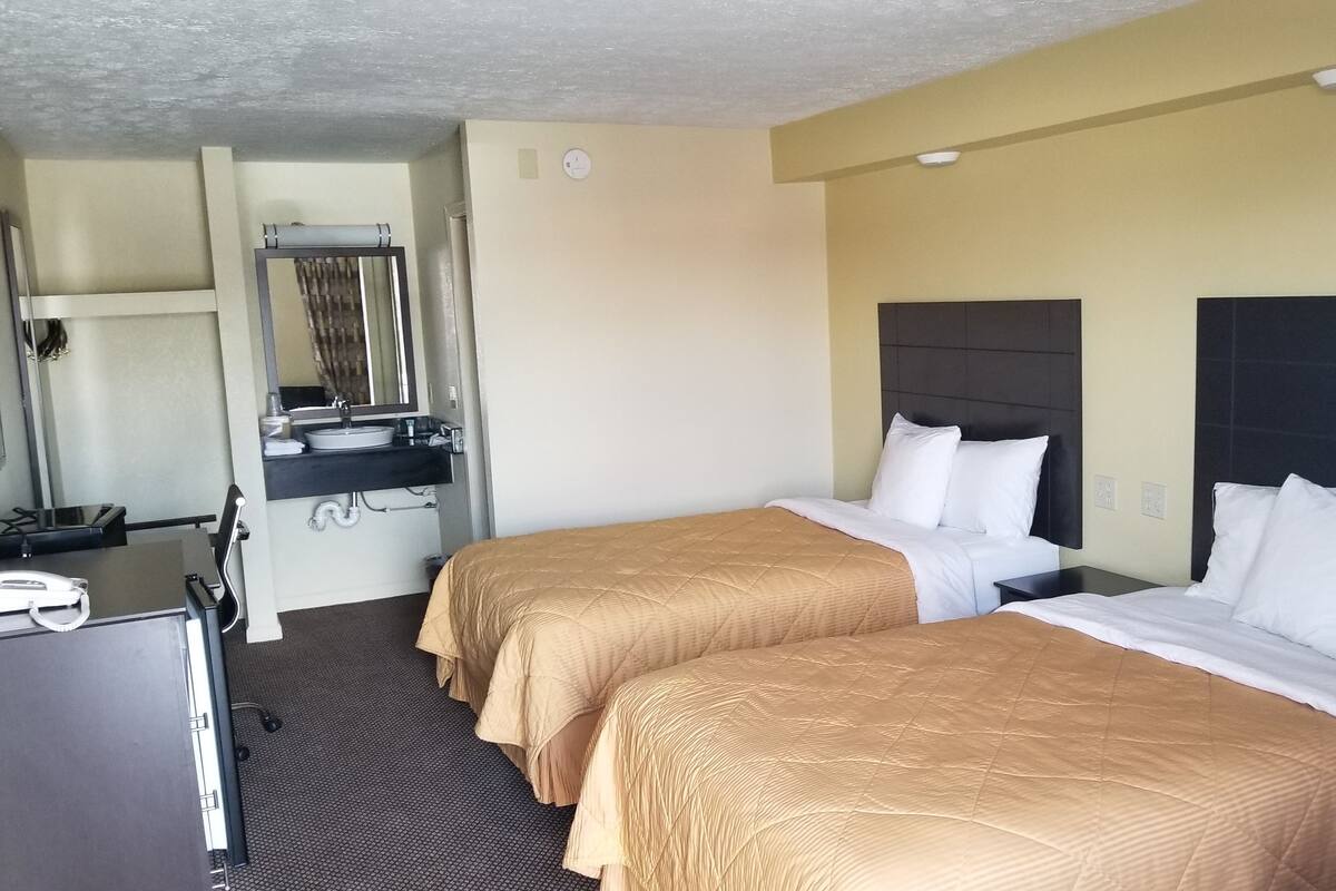 A hotel room with two beds, a desk, a mirror, and a sink.