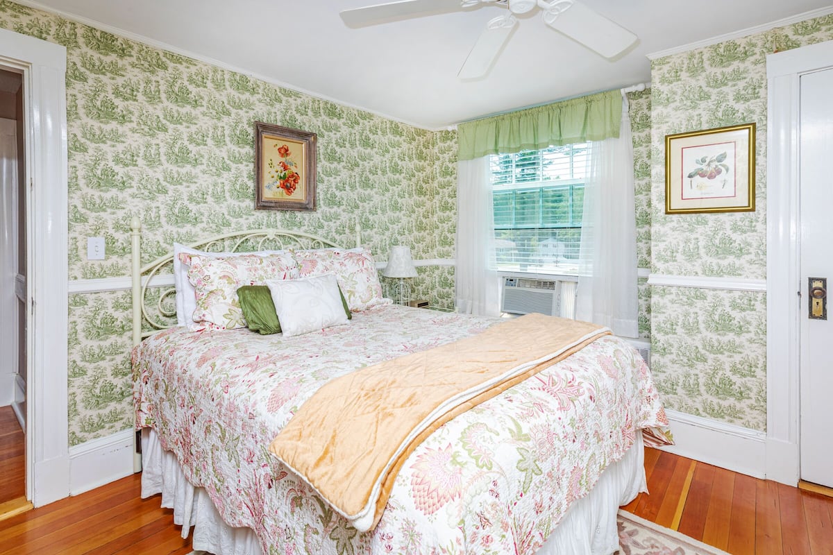A bedroom with floral wallpaper, a bed with a floral bedspread, a ceiling fan, and a window with blinds.
