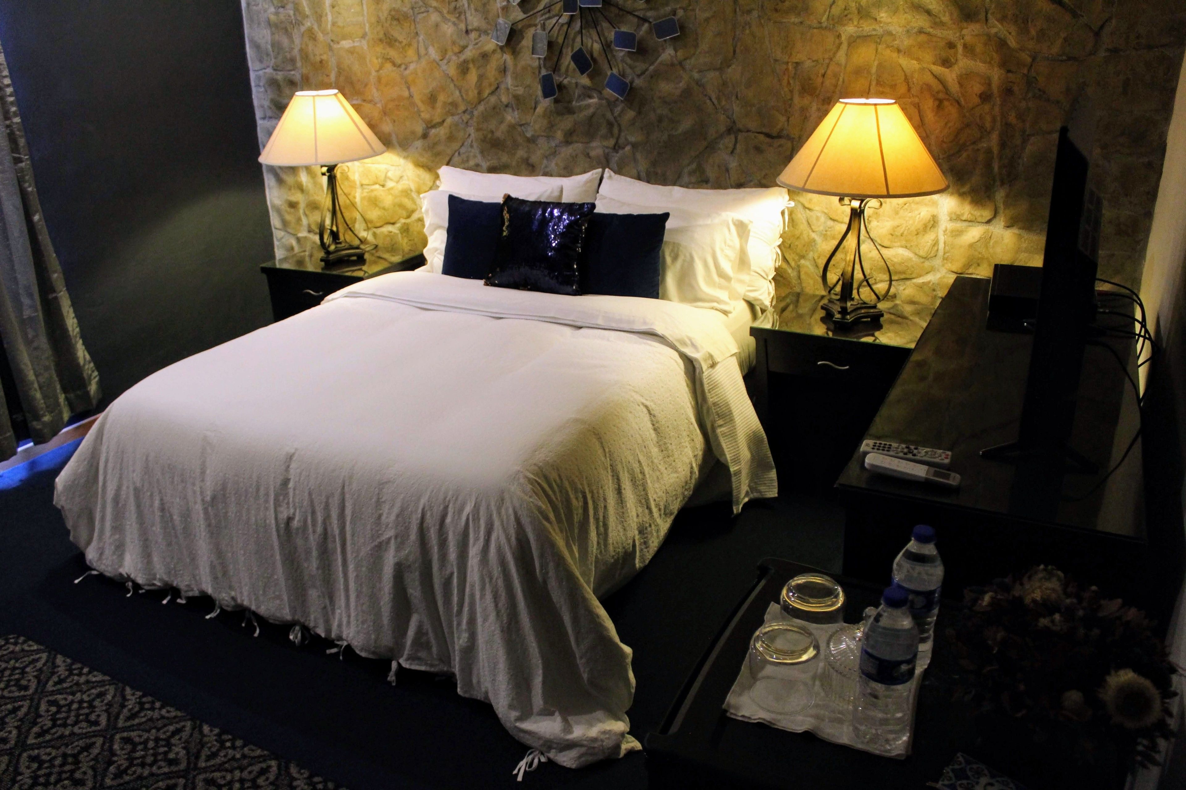 A neatly made bed with white linens, two bedside lamps, a stone wall, a small table with a remote control, and a tray with water bottles and glasses.
