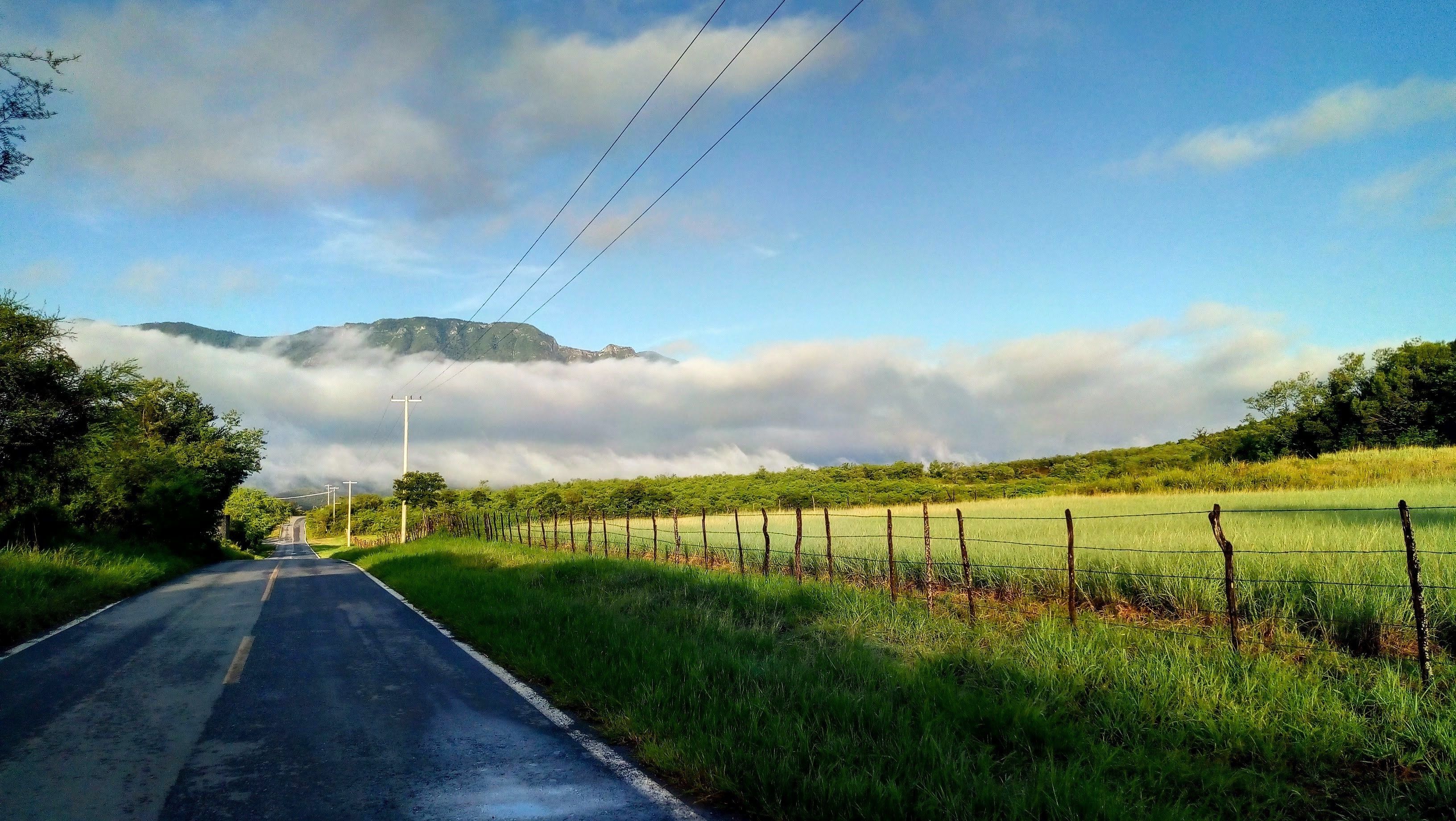 A rural road with a mountain in the background, surrounded by greenery and a fence.