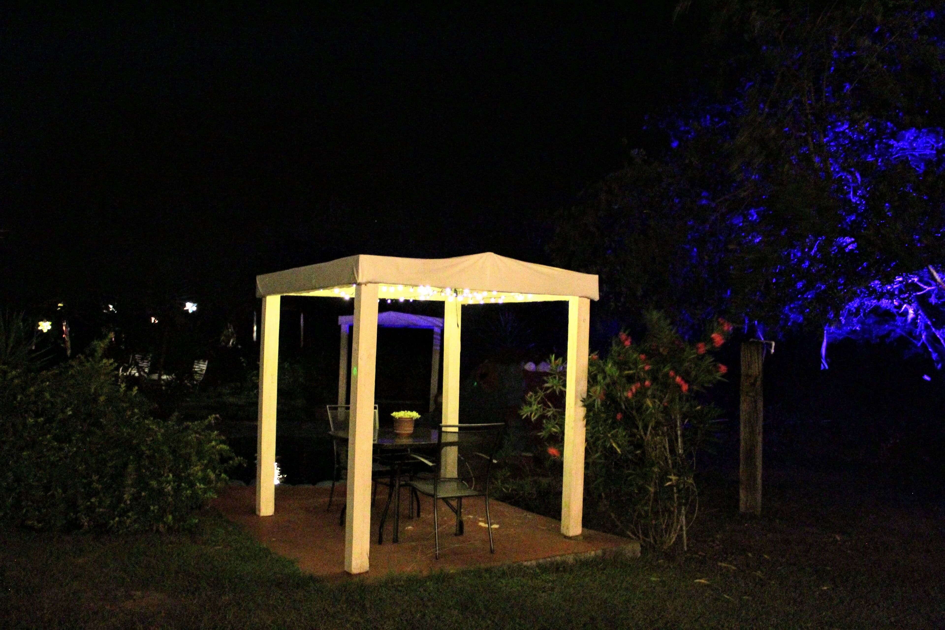 Een prieeltje met een tafel en stoelen, omringd door planten en bomen.