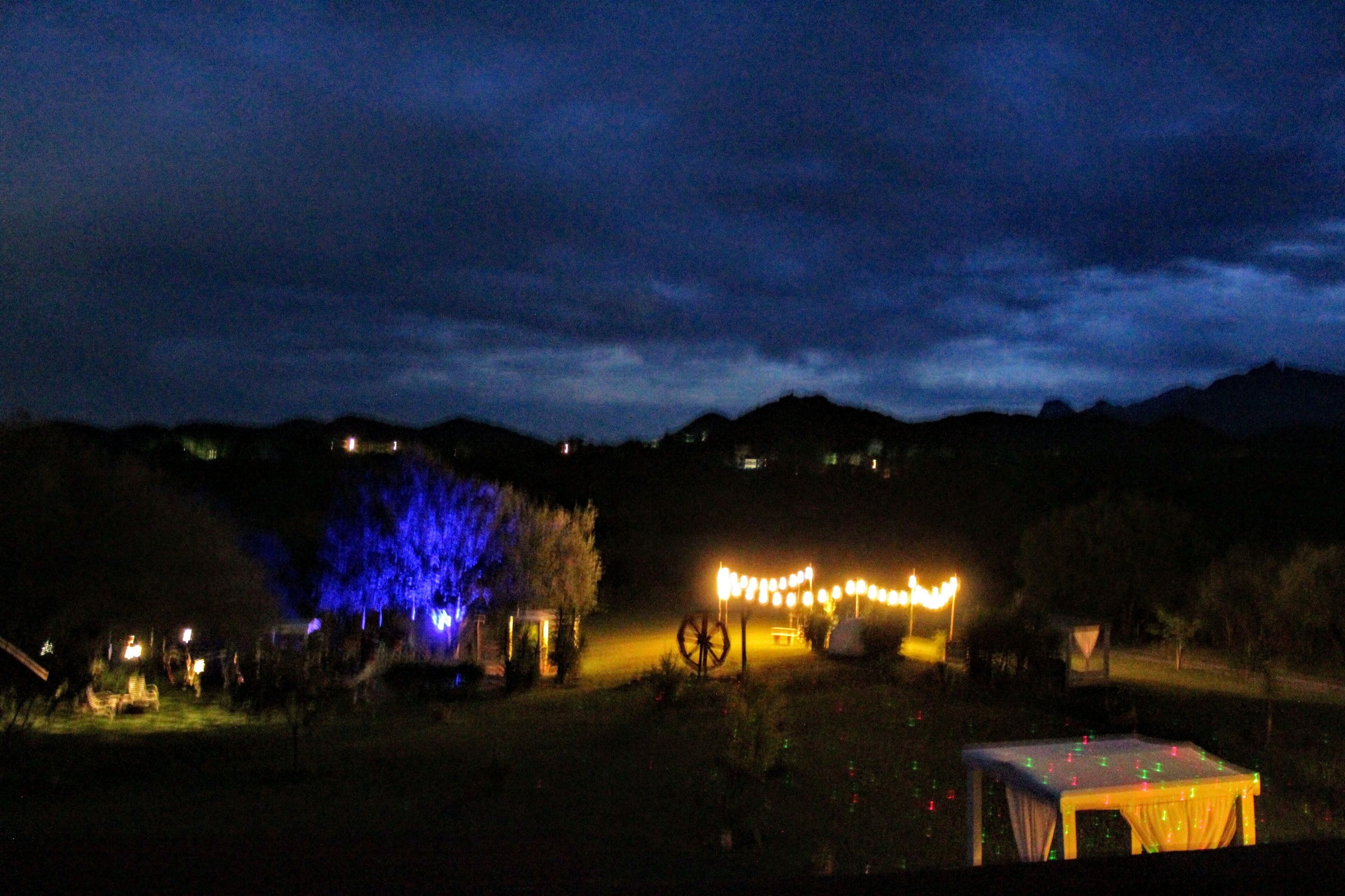 Vista nocturna de un paisaje rural con una pequeña estructura iluminada, posiblemente un porche, rodeada de árboles y una colina distante.