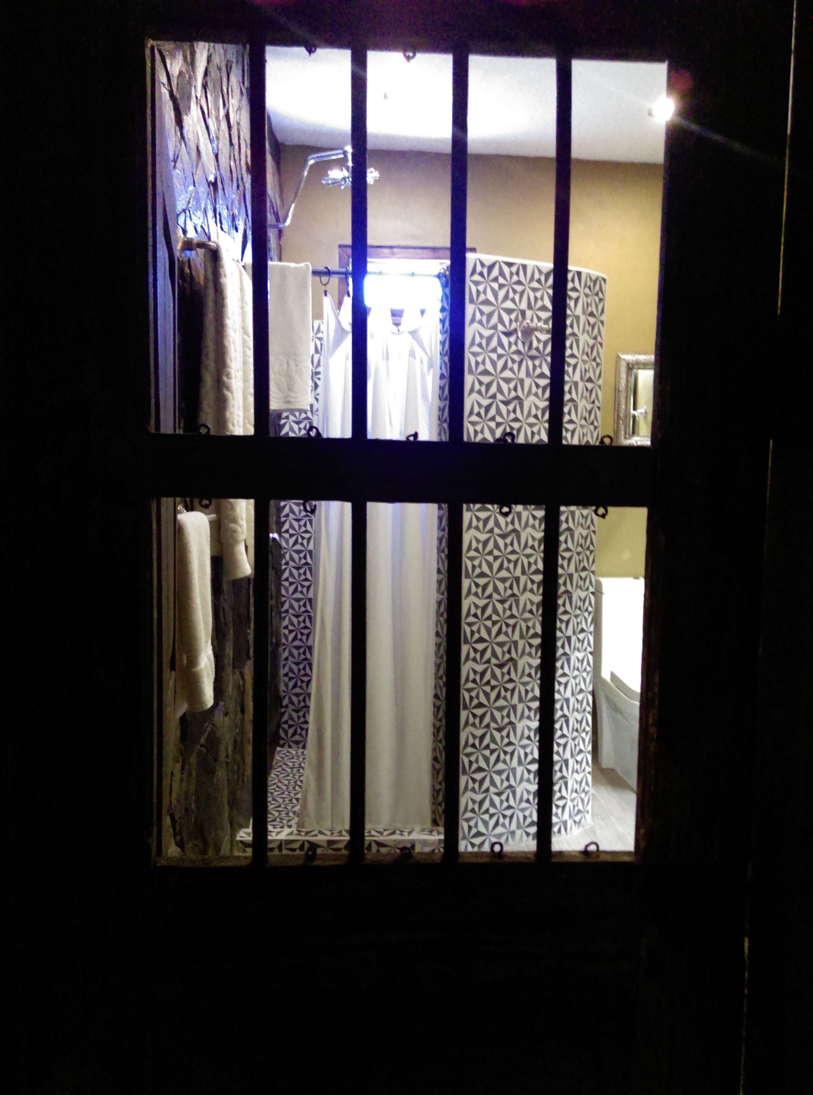 Vista a través de una ventana con barrotes que da a un baño con cortinas estampadas, un perchero y un espejo.