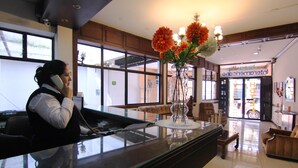 A hotel reception desk with a vase of flowers, a receptionist on the phone, and a view of the hotel entrance.