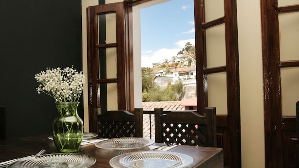 A dining table set with plates, a vase with flowers, and a view of a hillside through open windows.