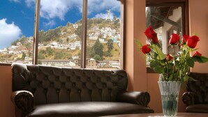 A room with a leather sofa, a round wooden table, and a vase with red roses. A large window offers a view of a hillside with buildings.