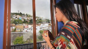 A person holding a mug, looking out of a window with a view of a hillside town.
