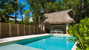 A swimming pool with a thatched-roof structure, surrounded by lush greenery and a wooden fence. - Tulum Attha (Tulum)