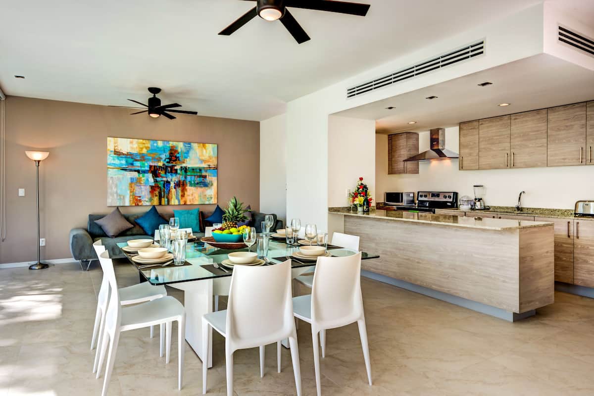 Una cocina moderna con una isla central, electrodomésticos integrados y un comedor con mesa de vidrio y sillas blancas.