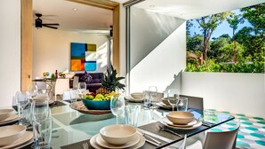 A dining area with a glass table, chairs, and a fruit bowl. There is a view of a garden through a large window. - Tulum Attha (Tulum)